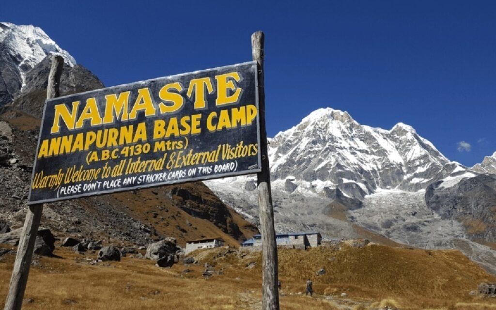 Close-up of a trekker standing at Annapurna Base Camp signboard with Annapurna I towering behind under a clear blue sky.