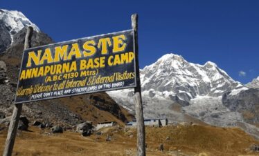 Close-up of a trekker standing at Annapurna Base Camp signboard with Annapurna I towering behind under a clear blue sky.