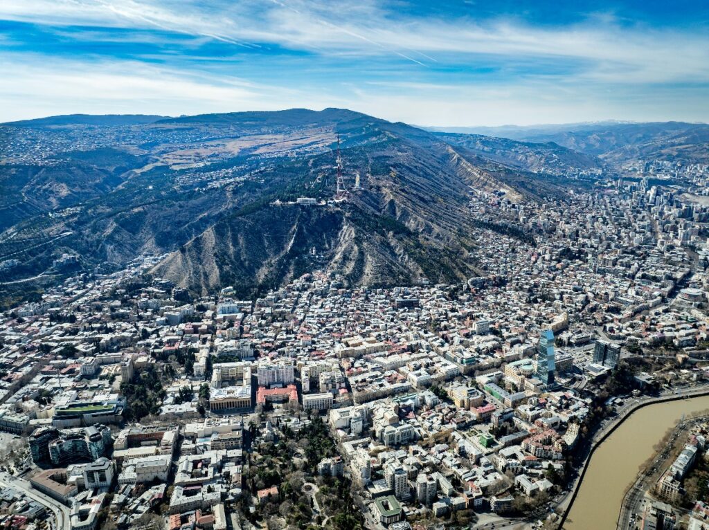 Panoramic aerial view of Tbilisi, Georgia showing Mtatsminda Hill, TV tower, and cityscape along the Kura River under a clear blue sky