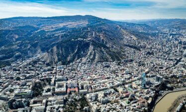 Panoramic aerial view of Tbilisi, Georgia showing Mtatsminda Hill, TV tower, and cityscape along the Kura River under a clear blue sky