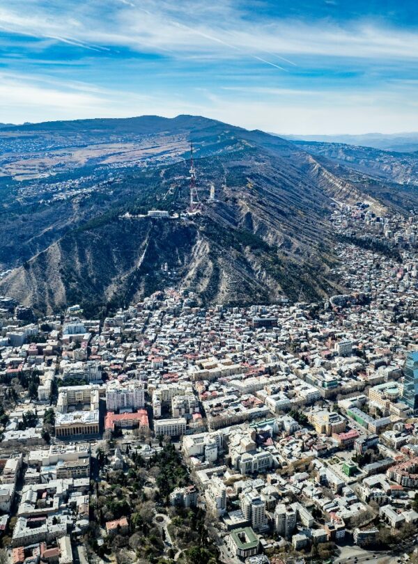 Panoramic aerial view of Tbilisi, Georgia showing Mtatsminda Hill, TV tower, and cityscape along the Kura River under a clear blue sky