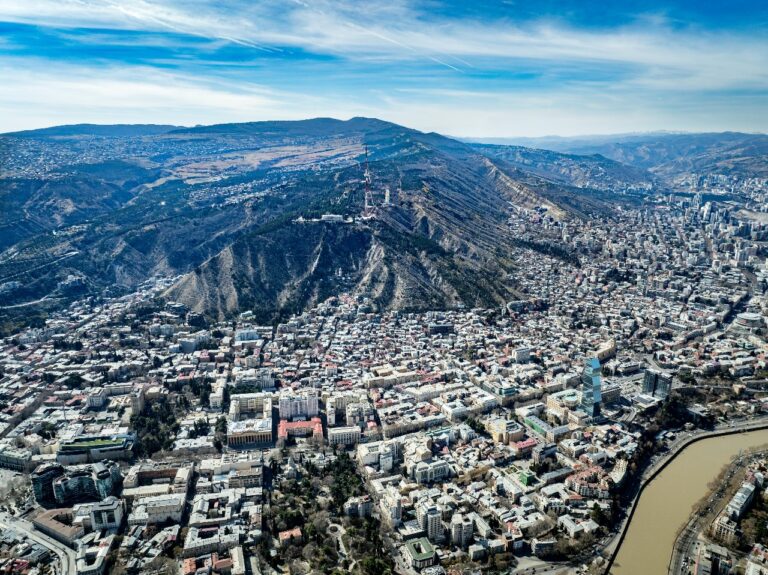 Panoramic aerial view of Tbilisi, Georgia showing Mtatsminda Hill, TV tower, and cityscape along the Kura River under a clear blue sky