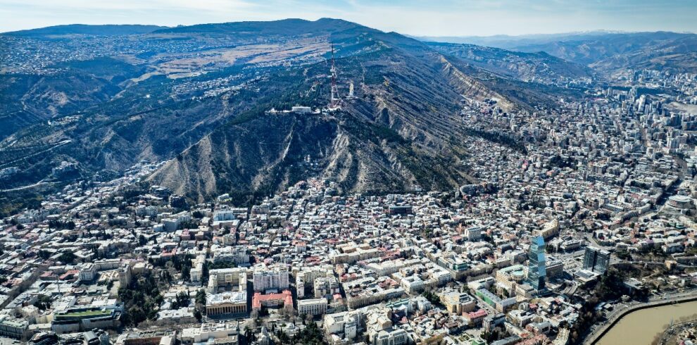 Panoramic aerial view of Tbilisi, Georgia showing Mtatsminda Hill, TV tower, and cityscape along the Kura River under a clear blue sky